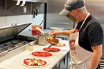 A person wearing a cap, face mask, and apron prepares pizzas in a commercial kitchen, spreading sauce onto dough lined up on a counter.