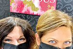 Two women wearing black face masks are standing side by side, smiling with their eyes visible. A pink and white abstract painting hangs on a patterned wall behind them.