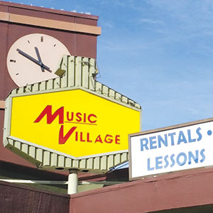 A bright yellow sign reading Music Village stands near a building with a large clock. Another sign nearby says Rentals • Lessons against a clear blue sky.