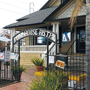 A tan house with black trim and a front porch has a sign reading “Roadhouse Bistro” over its entrance gate. An “OPEN” sign hangs by the steps, and various notices are posted on the gate and fence.