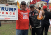 Two people at a rally, one holding a Trump Pence 2020 sign and the other wearing a Trump hat and holding a Trump paddle, surrounded by others with flags and pro-Trump signs.