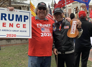 Two people at a rally, one holding a Trump Pence 2020 sign and the other wearing a Trump hat and holding a Trump paddle, surrounded by others with flags and pro-Trump signs.