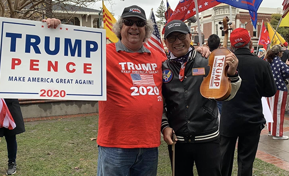 Two people at a rally, one holding a Trump Pence 2020 sign and the other wearing a Trump hat and holding a Trump paddle, surrounded by others with flags and pro-Trump signs.