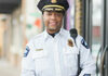 A police officer in uniform smiles while standing outside near a building entrance. He wears a hat and a radio on his shoulder.