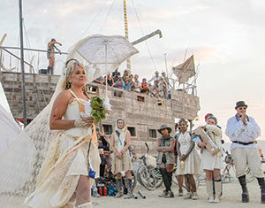 A woman in a white dress, winged cape, and boots holds a bouquet and parasol, walking past a group of people. Behind them is a large wooden ship structure with people aboard, set in a desert environment.