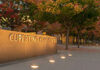 A stone sign with Cupertino City Center engraved on it is illuminated by ground lights, surrounded by trees with autumn foliage in a landscaped urban area.