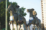 Two bronze statues of people riding horses are displayed outdoors, with trees and buildings in the background. Both statues have paper signs attached to their chests.