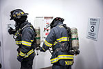 Two firefighters in full gear and breathing apparatus stand next to a fire safety cabinet on the third floor of a building, preparing for action.