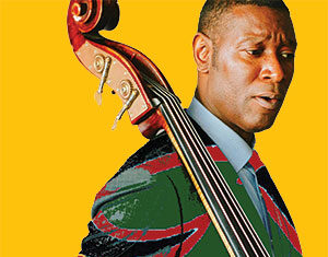 A man in a patterned suit holds an upright bass over his shoulder against a solid yellow background.