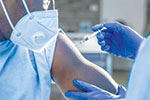 A healthcare worker wearing blue gloves prepares to administer an injection into a person’s arm. The person receiving the shot is wearing a face mask.