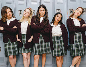 Five young women stand in front of blue lockers, wearing matching plaid skirts, white blouses, and maroon cardigans, posing confidently with arms crossed or hands on hips.