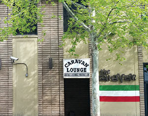 A beige brick building with a tree in front, a security camera, and a sign above a doorway that reads Caravan Lounge. The right side features green, white, and red stripes and faded letters on the wall.