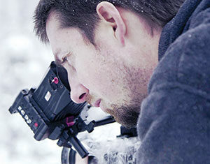 A man with a short beard in a dark jacket is closely looking through the viewfinder of a professional video camera while filming outdoors in a snowy environment.