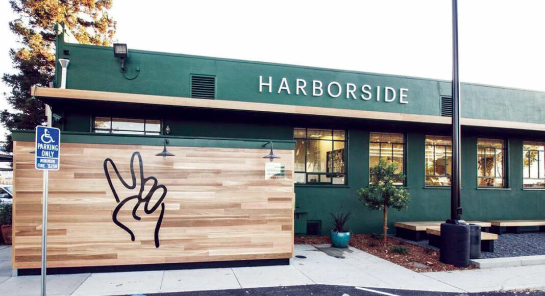 A green building with the sign Harborside above large windows, a wooden wall featuring a black peace hand sign, and a parking lot with a handicap parking sign in the foreground.