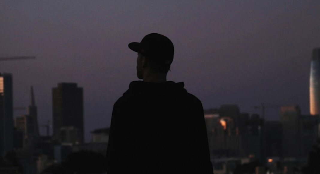 A person in a hoodie and cap stands with their back to the camera, silhouetted against a city skyline at dusk, with tall buildings and a crane visible in the background.
