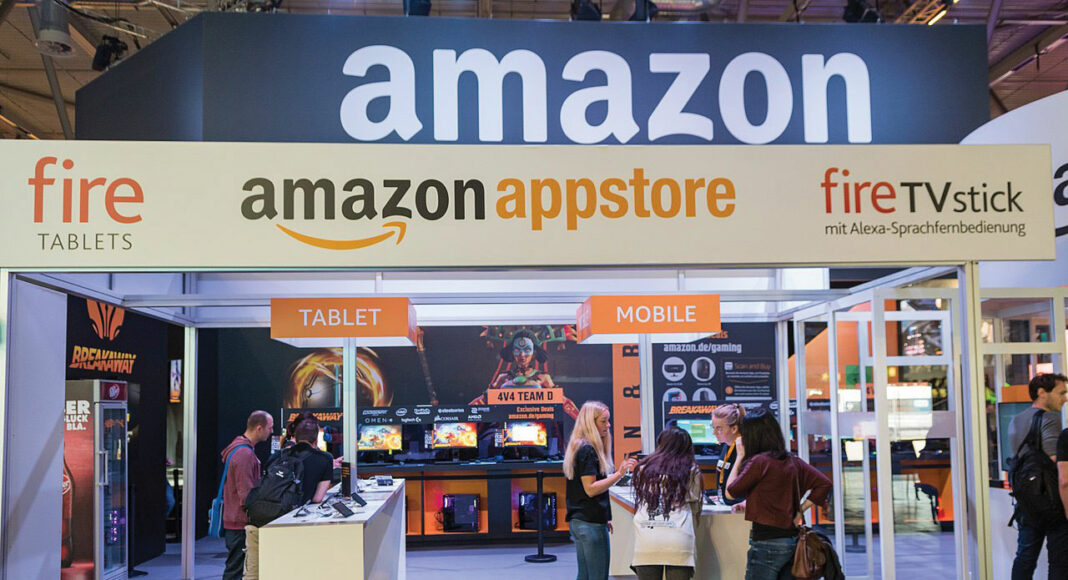 Amazon appstore booth at a tech event, with people interacting near display tables labeled ‘Tablet’ and ‘Mobile.’ Fire Tablets and Fire TV Stick banners are visible, and gaming screens are in the background.