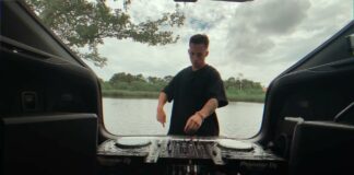 A person stands at the back of a vehicle by a river, using DJ equipment set up in the trunk, with trees and a cloudy sky in the background.