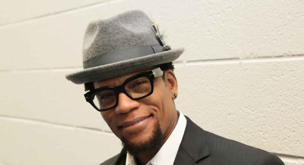 A man wearing a gray fedora, black glasses, a black suit, and a polka dot tie smiles while standing against a white brick wall. He has a neatly trimmed beard and a red pin on his lapel.