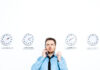 A man in a blue shirt and black tie looks confused while talking on the phone, standing in front of four clocks labeled Los Angeles, Wisconsin, Australia, and Galactic Wormhole, each showing different times.