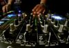 A person’s hands adjust controls and sliders on a professional DJ mixer, with illuminated buttons and colorful LED lights visible in a dimly lit setting.