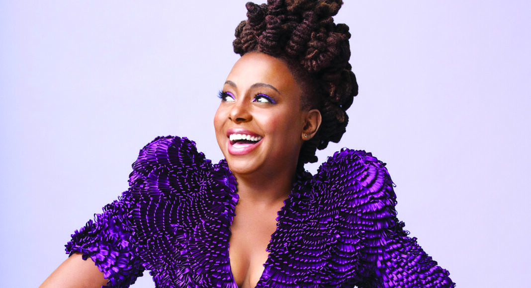 A woman smiles brightly while looking to the side. She wears a textured, deep purple dress with puffy sleeves and has her hair styled in an intricate updo. The background is a soft, light purple.