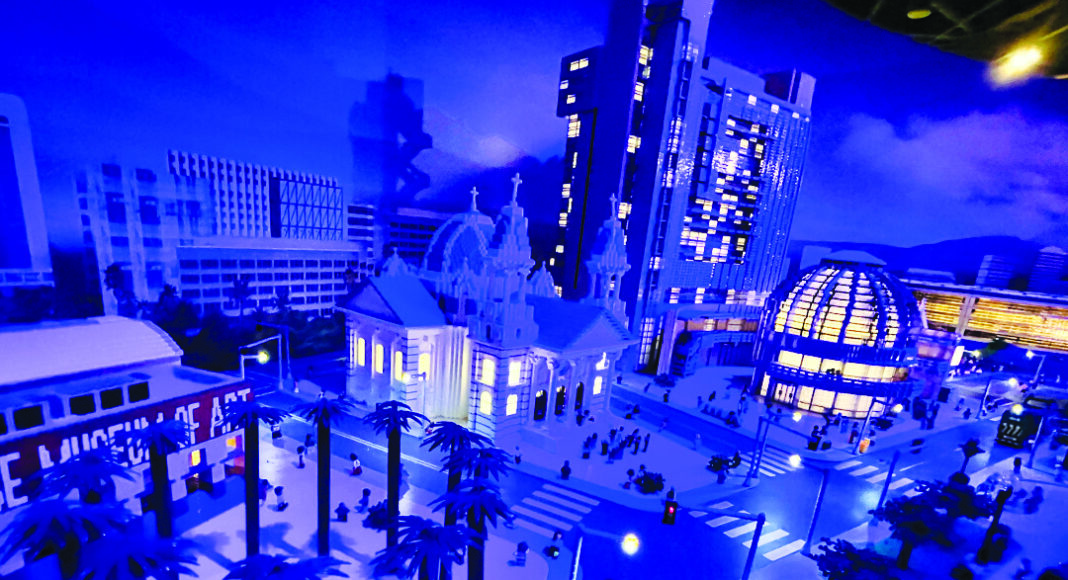 A miniature cityscape model features palm trees, a domed building, a church, high-rise buildings, and tiny people, all illuminated with blue and purple lighting for a vibrant, nighttime effect.