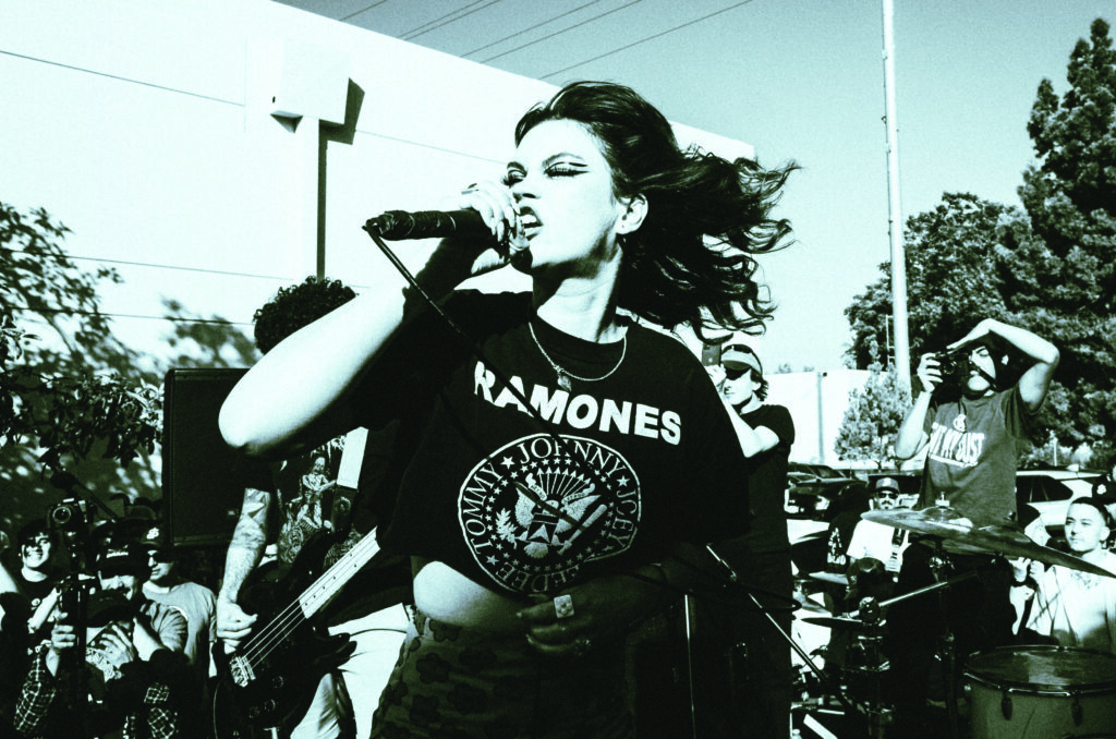 A woman in a Ramones T-shirt passionately sings into a microphone at an outdoor concert, surrounded by an energetic crowd and band members under bright daylight.