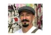 A man with a gray flat cap, curled mustache, and goatee smiles in front of a wall covered in colorful graffiti. He is wearing a white shirt and a brown strap over his shoulder.