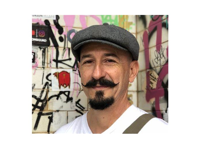 A man with a gray flat cap, curled mustache, and goatee smiles in front of a wall covered in colorful graffiti. He is wearing a white shirt and a brown strap over his shoulder.