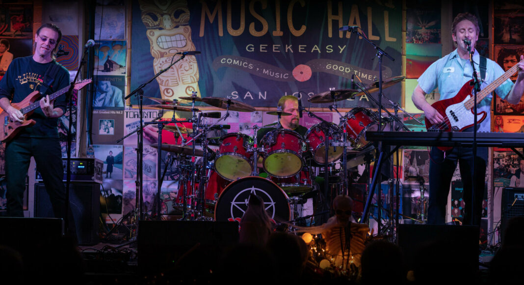 Three musicians perform on stage at Music Hall Geek Easy in San Jose. The guitarist, drummer, and bassist are playing under colorful lights, with posters and a large sign visible in the background.