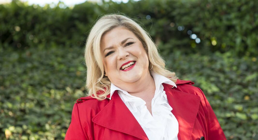 A smiling woman with blonde hair wearing a bright red coat over a white ruffled blouse, standing outdoors in front of green foliage.