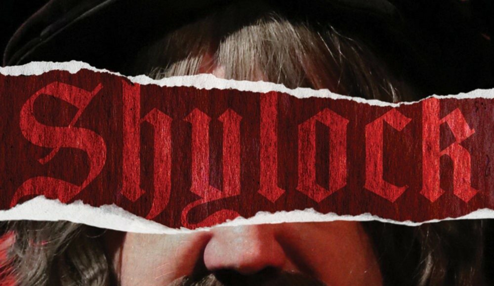 A close-up of a person with a gray beard and long hair, partially obscured by a torn piece of paper displaying the word Shylock in bold red letters.