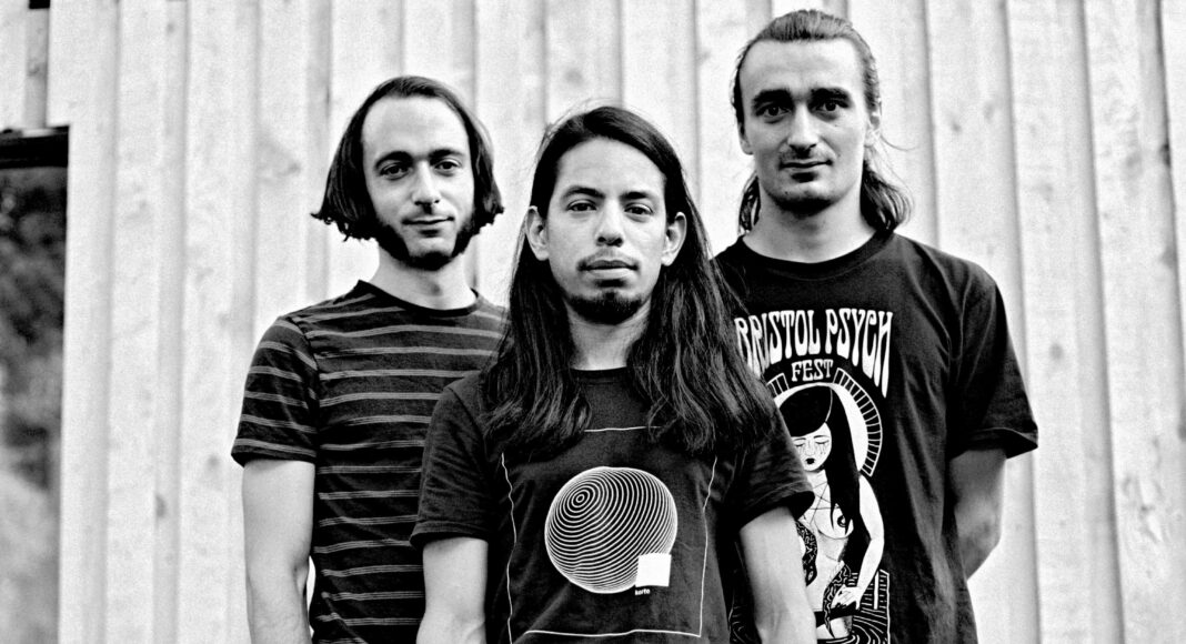 Three young men stand side by side in front of a wooden wall, facing the camera. All have shoulder-length hair and wear printed t-shirts. The photo is black and white.