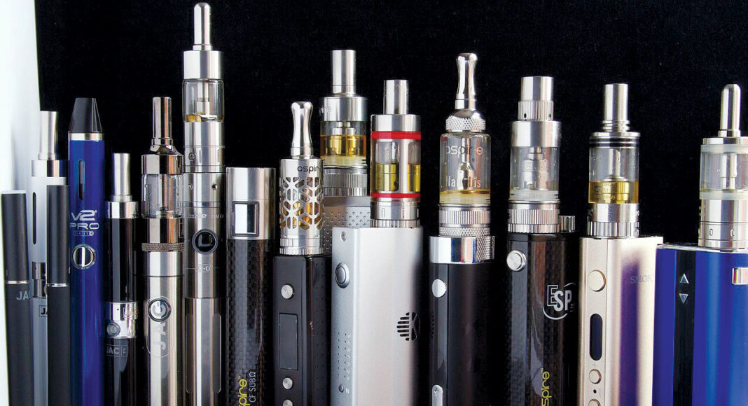 A variety of electronic vaping devices and vape pens, in different shapes, sizes, and colors, are arranged in a row against a black background.