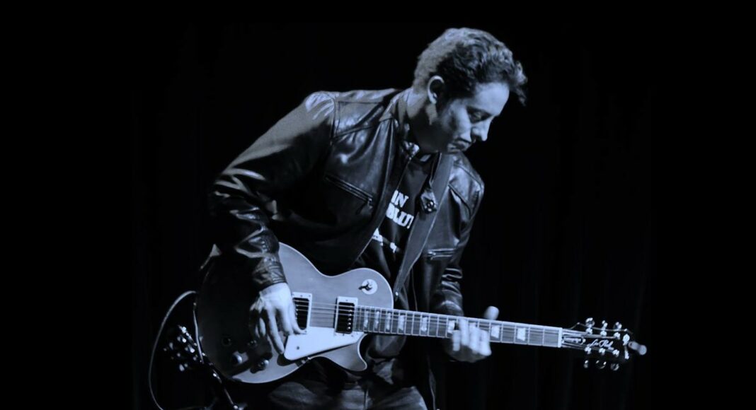 A man wearing a leather jacket plays an electric guitar on stage, with dark curtains in the background, under low lighting.
