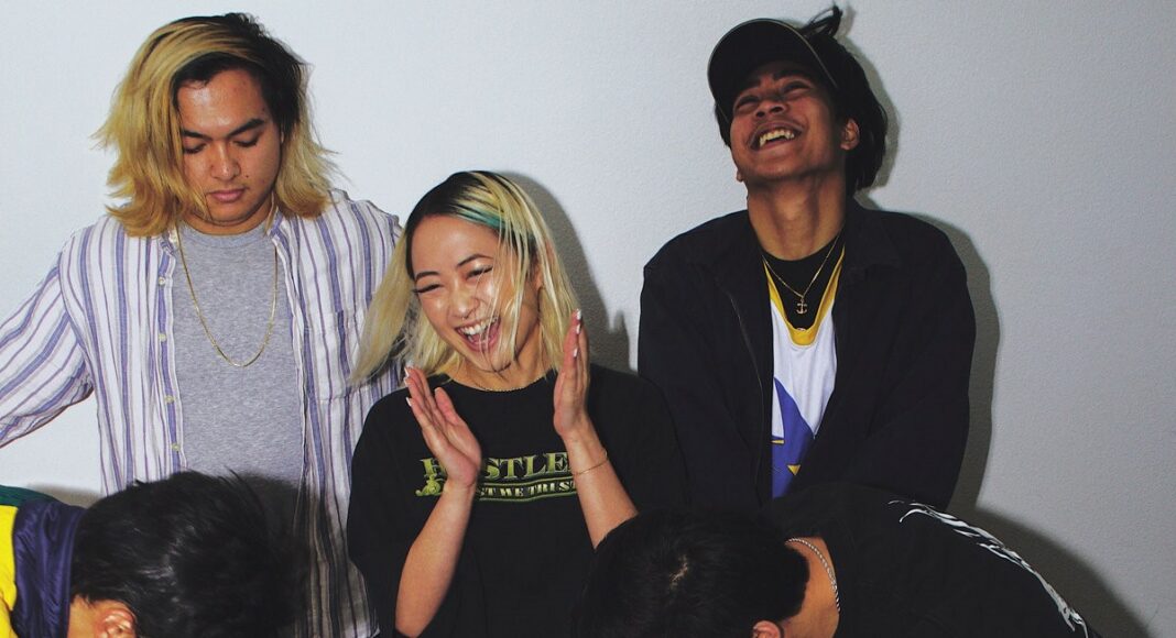 Five young adults stand against a plain white wall, laughing and smiling. One person claps, two look down, and two look up grinning, creating a joyful, candid, and playful atmosphere.