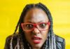 A person with braided hair, wearing red glasses and a black jacket, looks directly at the camera with a confused or surprised expression. The background is bright yellow.