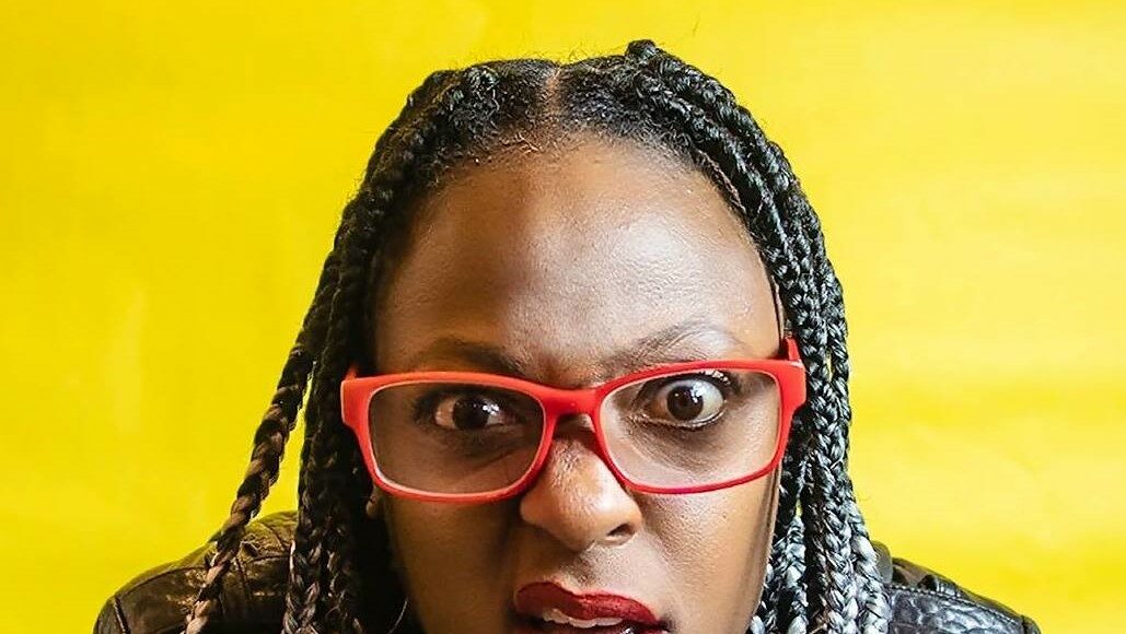 A person with braided hair, wearing red glasses and a black jacket, looks directly at the camera with a confused or surprised expression. The background is bright yellow.