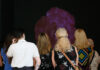A group of people stands facing a large purple flower displayed on a dark background, their backs turned to the camera as they observe the art closely.