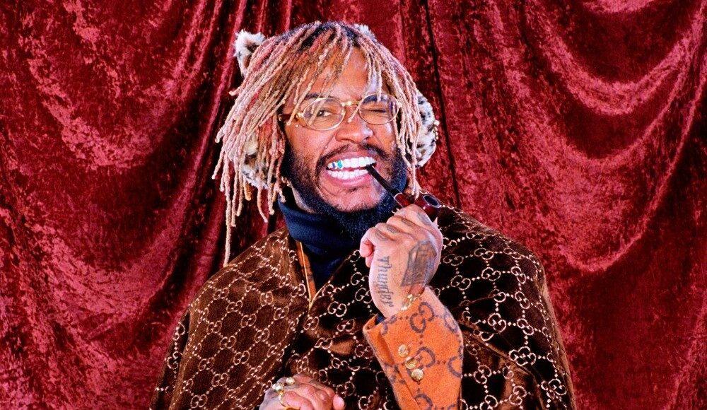 A smiling man with blond dreadlocks holds a pipe and wears glasses, a brown Gucci patterned coat, and rings. He stands in front of a red velvet curtain.