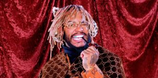 A smiling man with blond dreadlocks holds a pipe and wears glasses, a brown Gucci patterned coat, and rings. He stands in front of a red velvet curtain.