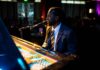 A man in a suit plays a grand piano and sings into a microphone on stage in a dimly lit venue with an audience in the background.