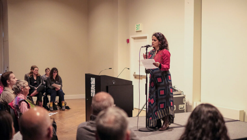 A woman in a patterned skirt speaks into a microphone on a small stage, holding papers. An audience sits facing her, listening attentively in a softly lit room. A podium is visible to the left.
