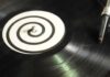 A close-up of a spinning vinyl record on a turntable, with a black-and-white spiral pattern on the label and the needle arm positioned on the record’s outer edge.
