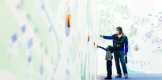 An adult and a child, both wearing masks, interact with buttons on a brightly lit, illustrated wall display in an indoor setting. The adult points at the wall while the child reaches up to press a button.