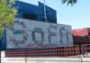 A mural spells out SOFA in colorful, abstract designs on a white wall outside a blue building, with a red railing and a tree in the foreground.