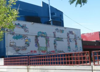 A mural spells out SOFA in colorful, abstract designs on a white wall outside a blue building, with a red railing and a tree in the foreground.