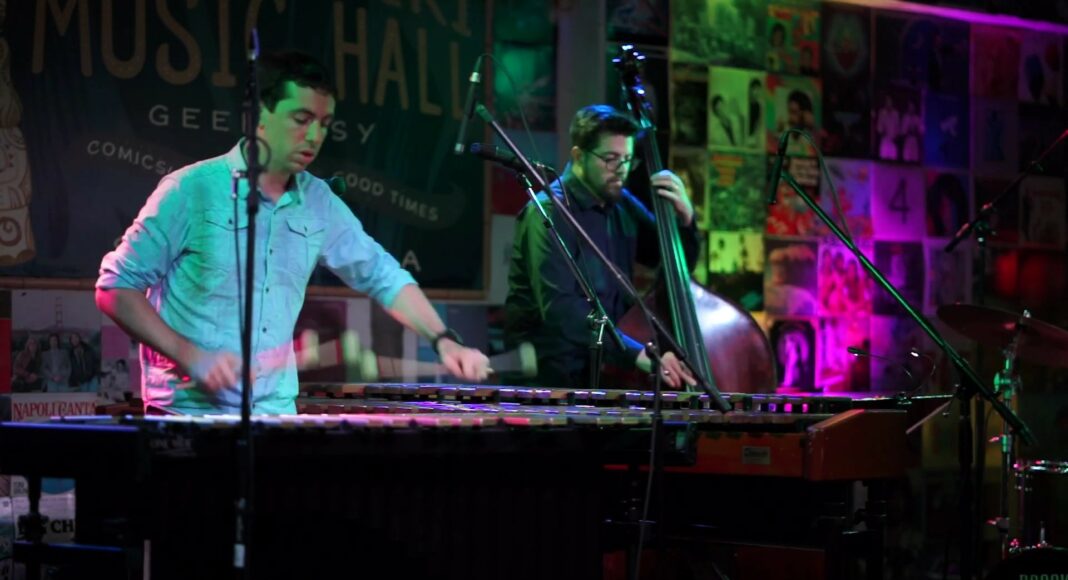 A musician plays a vibraphone while another plays an upright bass on a colorful stage decorated with posters and neon lights, with microphones set up around them.