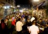 A lively outdoor night scene shows a crowd of people socializing around tall tables on a city street decorated with string lights and a rainbow flag. The atmosphere is festive and energetic.