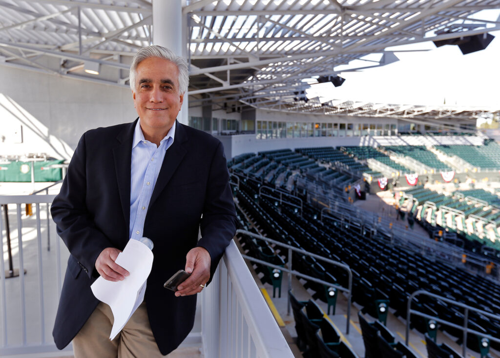 A man in a dark blazer and light pants stands smiling while holding papers and a phone in a mostly empty outdoor stadium with rows of green seats and a metal roof structure.